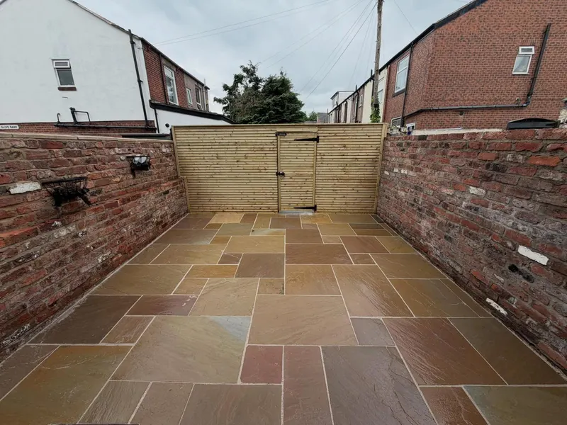 Warm-toned mixed-size natural stone flags in a long rectangular yard between brick walls, facing a light horizontal-slat gate and fence.
