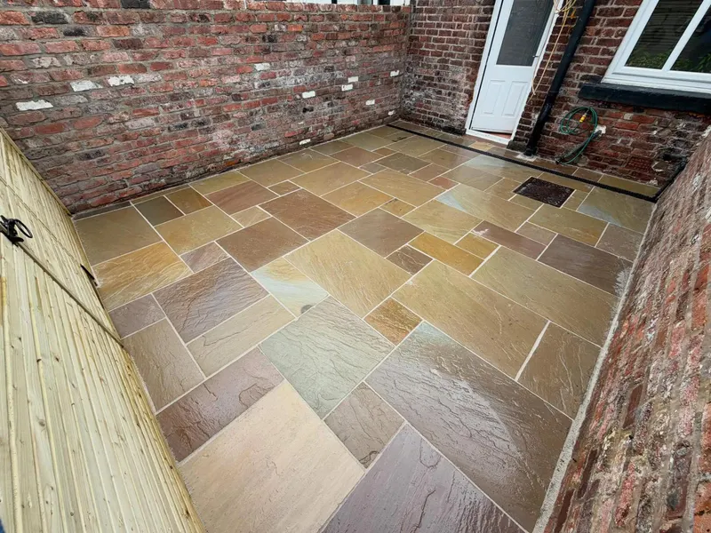 Multi-toned riven stone slabs in a small courtyard of aged brick walls and a timber fence, with channel drainage and an inset cover.