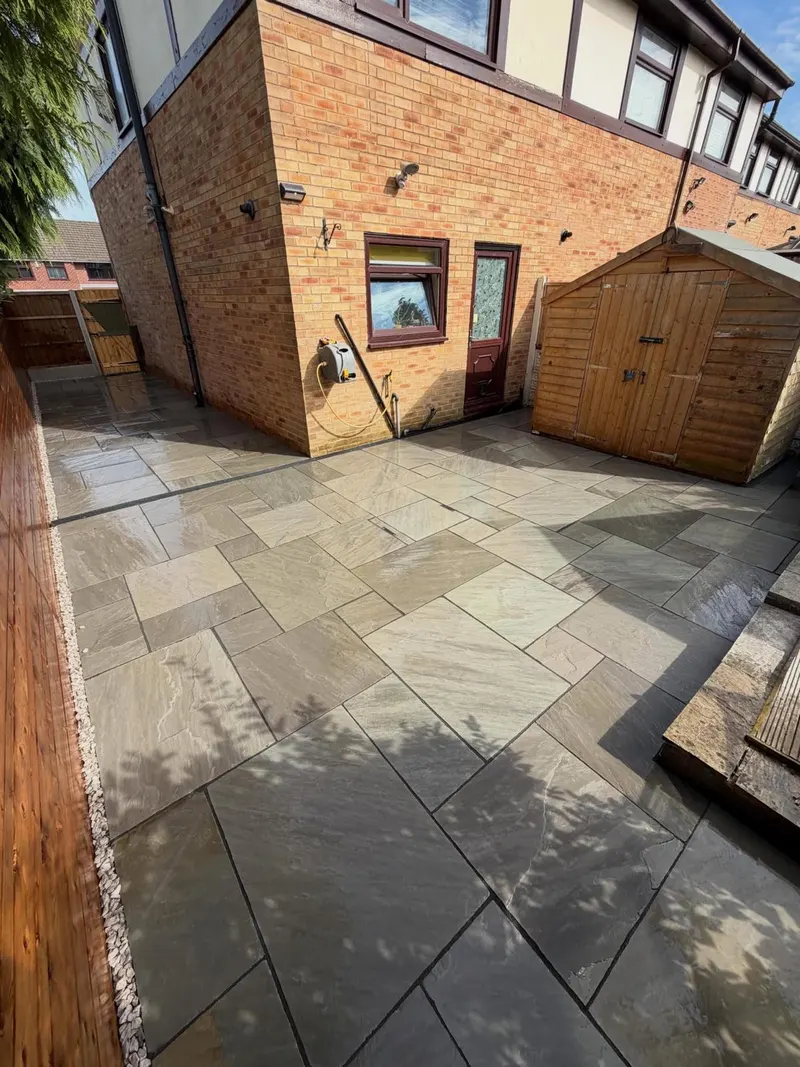 Light mixed natural stone flags across a back patio and narrow side path, with white gravel beside a fence, a shed, and a cross drain.
