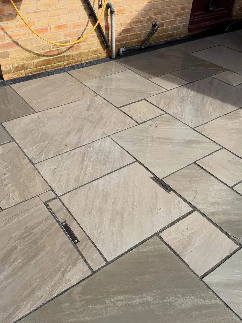Close-up of beige and tan riven stone paving with dark joints, showing a recessed access cover paved to match the surrounding pattern.