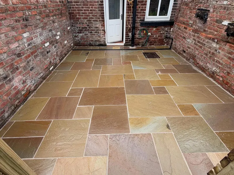 Multi-coloured riven stone slabs in a brick-walled courtyard, with a channel drain, inset utility cover, and white back door.