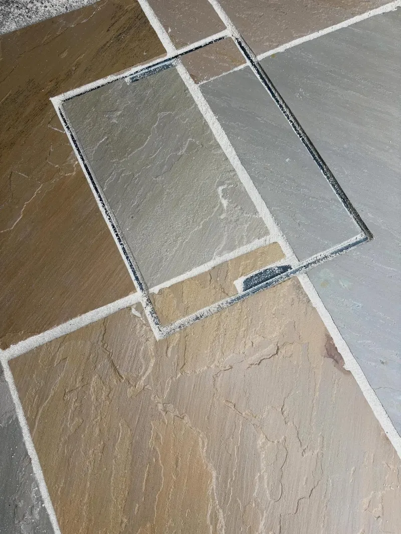 Detail of brown, tan, and grey riven sandstone slabs continuing through a thin-framed recessed manhole cover with light sand joints.