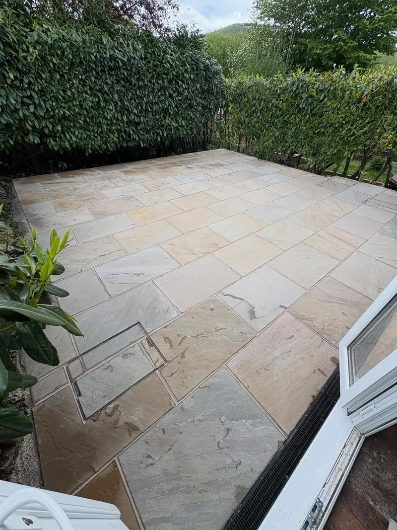 Tan and grey rectangular stone flags beside a tall hedge, with a paved-over recessed cover and a channel drain by a white door frame.