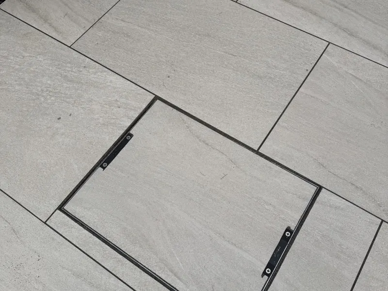 Light grey rectangular patio tiles surrounding a recessed metal access frame infilled with matching slabs and small lifting handles.