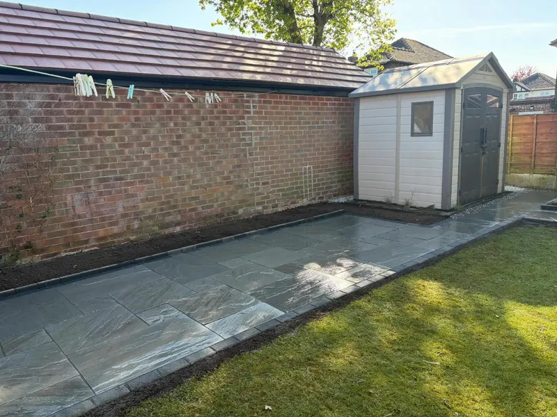 Grey veined stone flags in random layout with a charcoal block border, running beside a brick wall toward a garden shed and lawn.