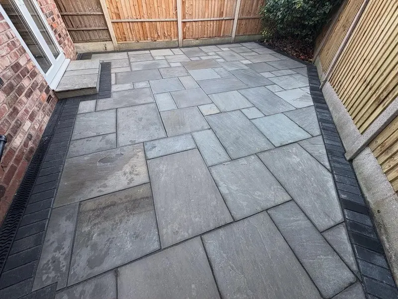 Mixed grey stone slabs enclosed by dark block edging, with a small paved step to glass doors, slot drain, and timber fencing.