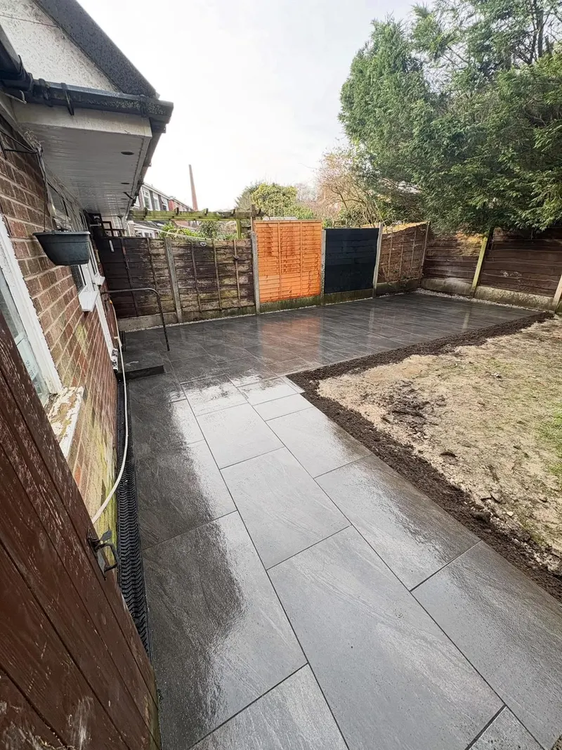 Large grey rectangular paving wrapping a brick house corner, with a channel drain at the wall and fresh soil beside the new surface.