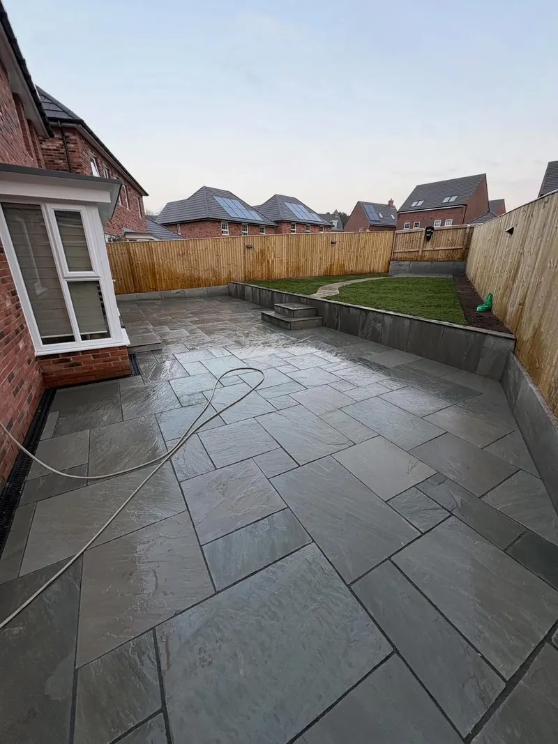 Multi-level grey stone flags with steps and a stone-clad retaining face, beside a brick bay window, lawn, and timber fence.