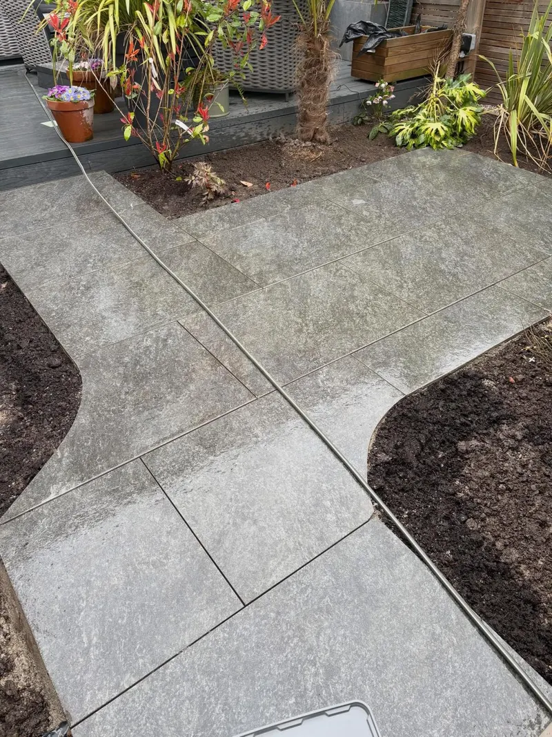 Large grey textured patio slabs forming a walkway that widens into a patio, with neat curved cuts at the corners beside fresh soil beds.