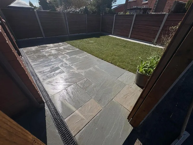Mixed grey and buff riven stone flags next to a rectangular new lawn, with a slot drain along the house and dark horizontal fencing.