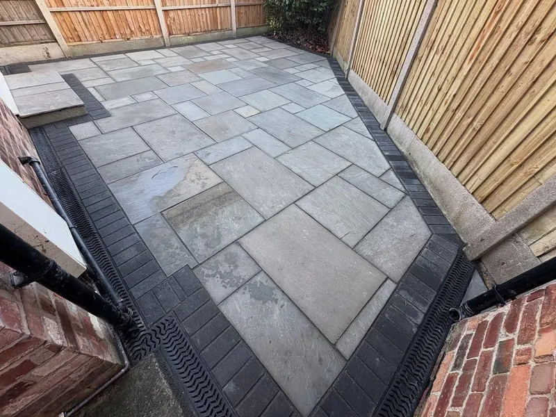 Light grey mixed-size stone patio framed by charcoal block edging and channel drains, with a small stone step and timber fencing.