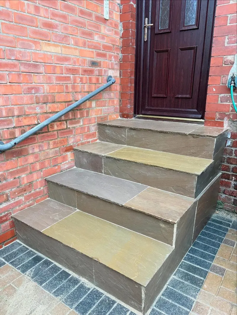 Four riven sandstone treads and risers with a dark block-paved landing and border, leading to a dark panelled door on red brick.