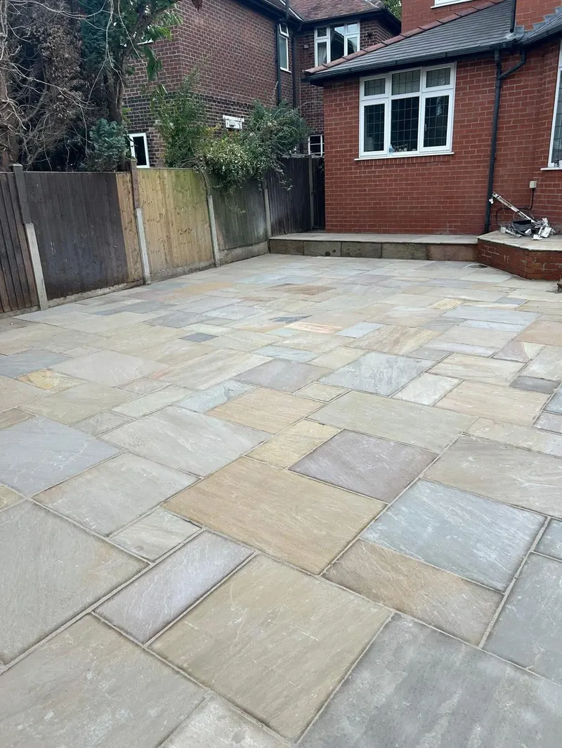 Large backyard paved in varied rectangular stone slabs in grey, buff and tan tones, laid beside a red brick house and wooden fence.