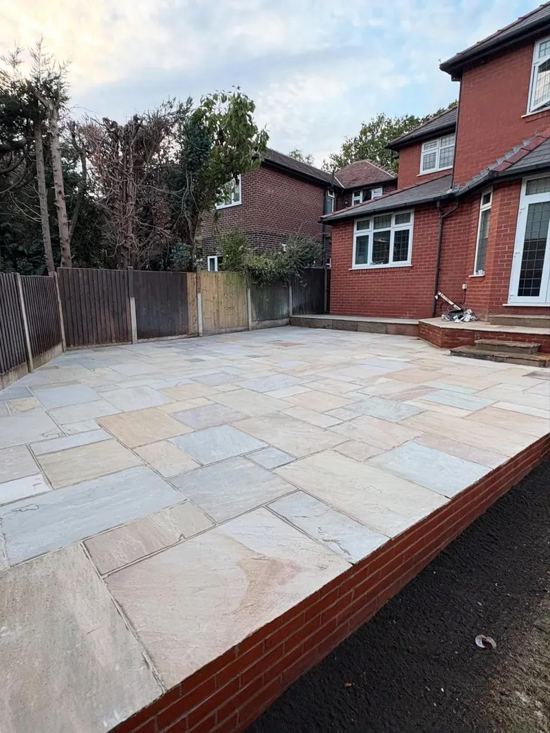 Raised stone patio with random-size flags, a red brick retaining edge and small steps leading to glass doors, with fresh soil still visible nearby.