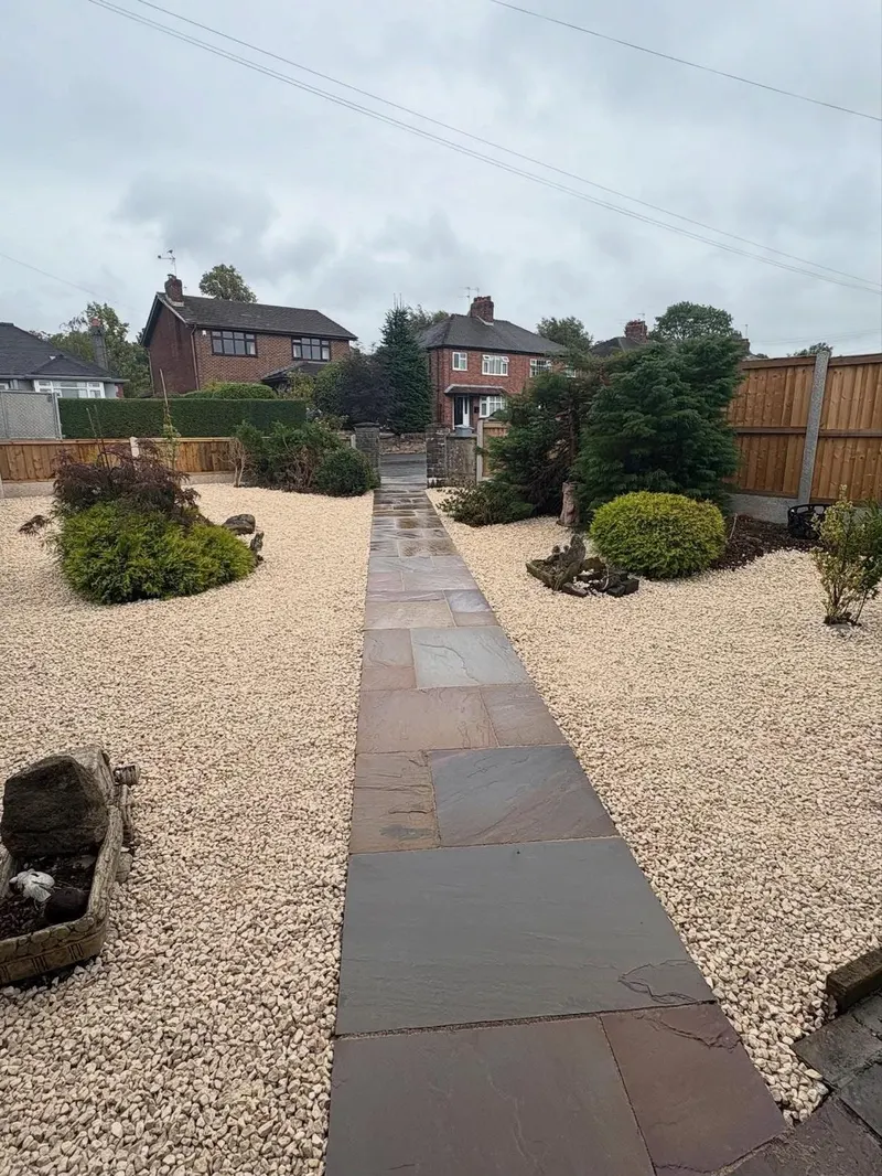 Straight garden path of large rectangular stone flags set in cream gravel, flanked by shrubs and decorative rocks in a suburban front garden.
