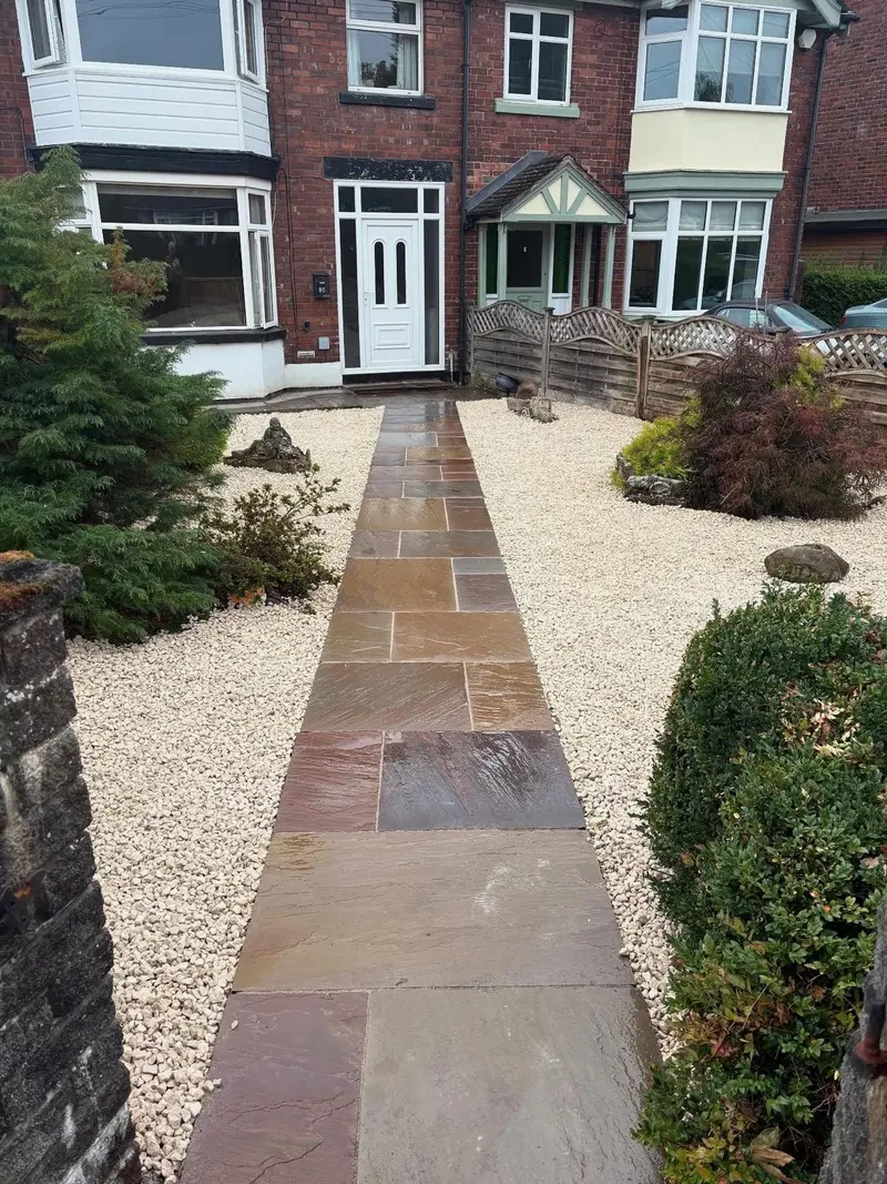 Narrow run of staggered riven stone slabs through pale gravel toward a white front door on a red brick semi-detached house.