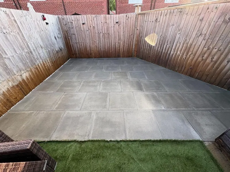 Square light grey paving slabs in a grid layout next to bright artificial grass, enclosed by tall wooden panel fencing.