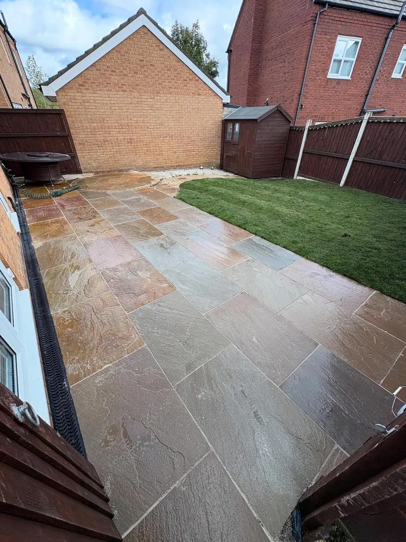 Riven stone flags in mixed buff and grey tones sweeping in a curved line against fresh green lawn, with a channel drain along the house wall.