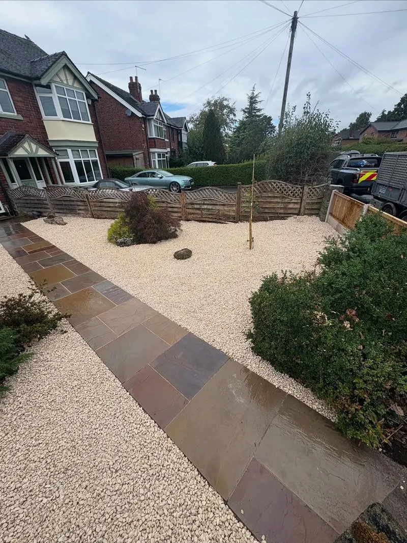 Indian sandstone slabs forming a straight path across a cream shingle front garden, with planting pockets and brick houses beyond the fence.