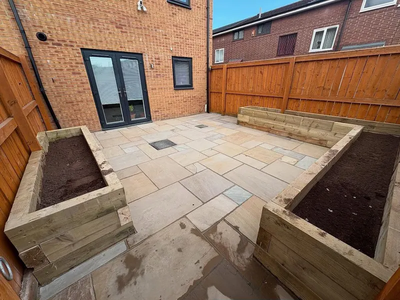 Back garden paved in mixed-size buff stone slabs surrounded by U-shaped raised beds built from chunky timber sleepers against a wooden fence.