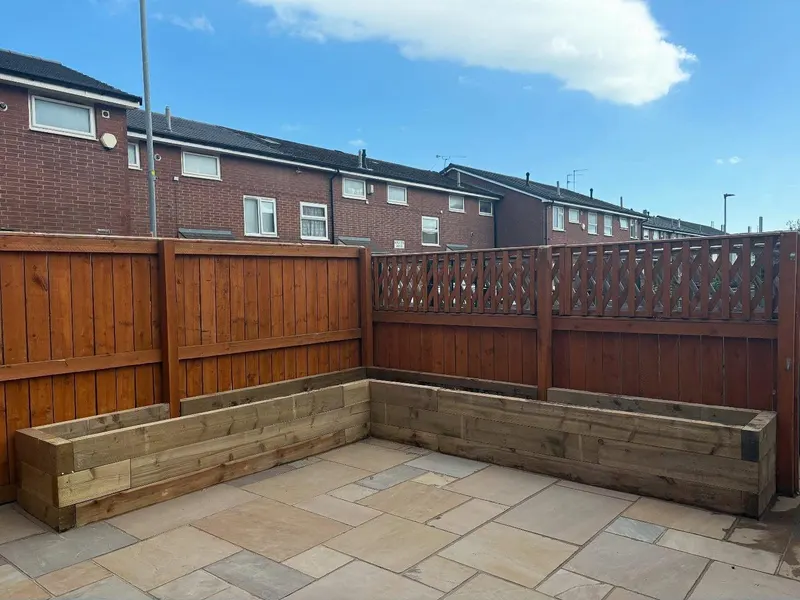 Beige stone slabs in a modular layout with an empty L-shaped timber sleeper planter in the corner, bounded by a brown stained fence.