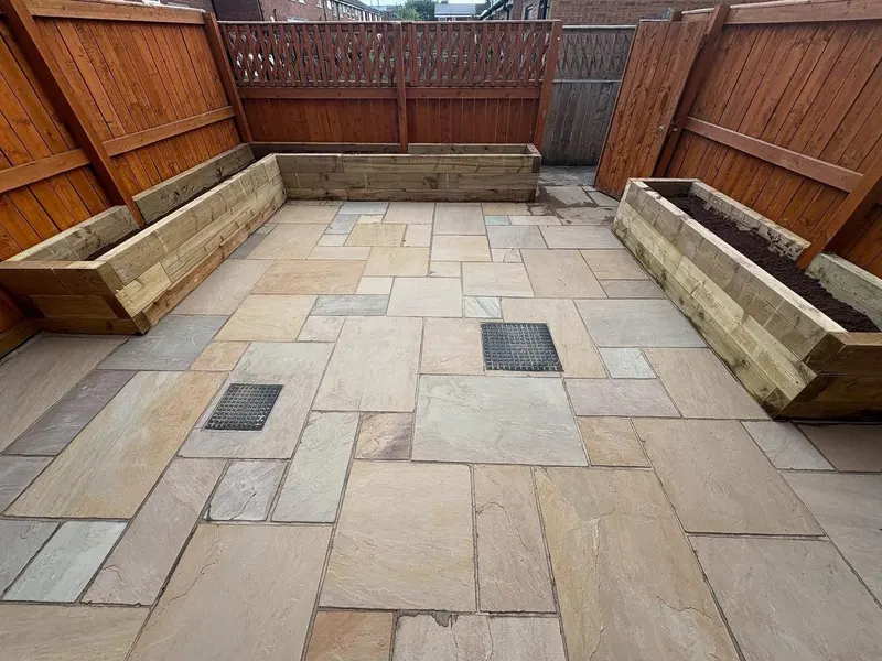 Overhead view of a fenced yard with multi-tonal flagstones, several timber sleeper planters and two flush black utility covers in the paving.