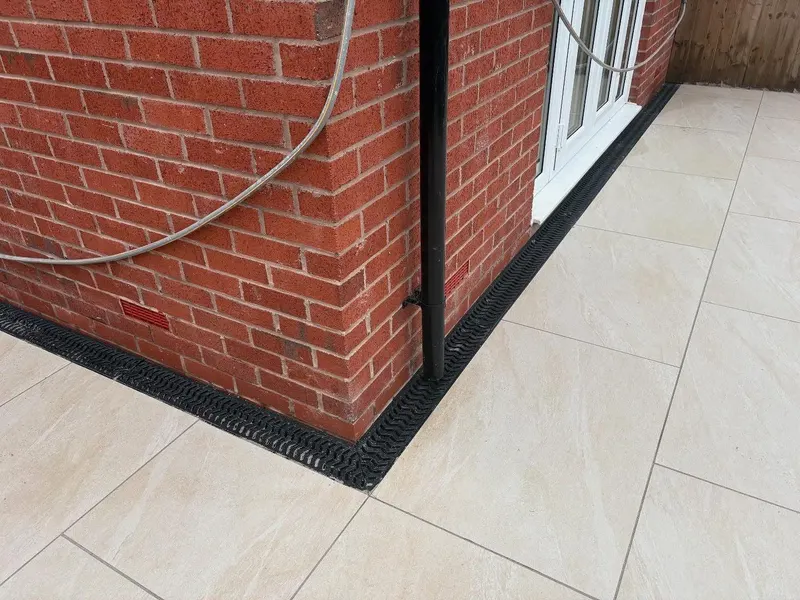 Close view of large light cream pavers meeting a red brick corner, with a black slot drain running along the wall below a downpipe.
