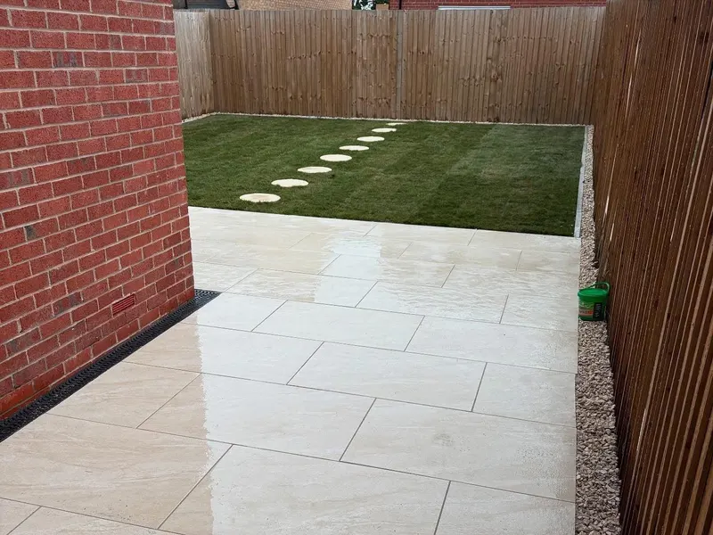 Light rectangular patio tiles beside a green lawn, linked by a row of round stepping stones toward the back corner of a fenced garden.