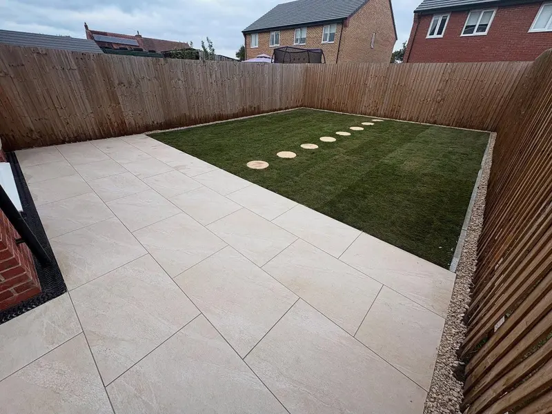 Wide light-beige tiled patio next to artificial grass, with seven circular stepping stones crossing the lawn diagonally to the fence.