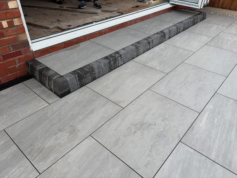 Large light grey slabs laid to bi-fold doors, framed by a raised border of small dark textured bricks forming a shallow entrance step.