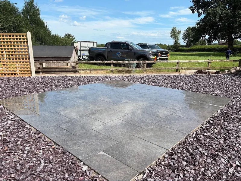 Square arrangement of smooth grey paving slabs surrounded by purple slate aggregate, with fencing and open fields in the background.