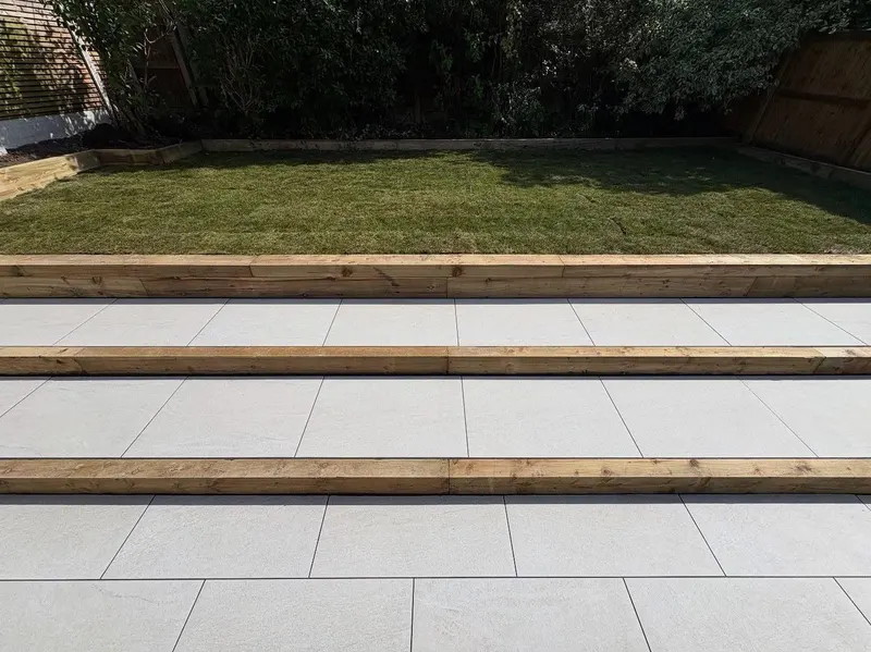 Three shallow wide steps with light grey tile treads and timber sleeper risers leading from paving up to a rectangular grass area.
