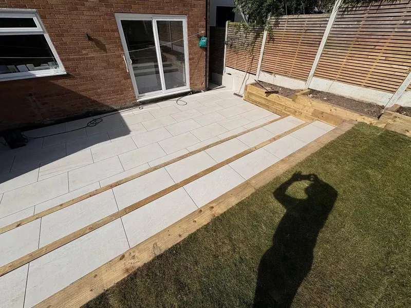 Light grey rectangular paving on two levels with timber sleeper step edges, beside a red brick house, lawn and a sleeper planter.