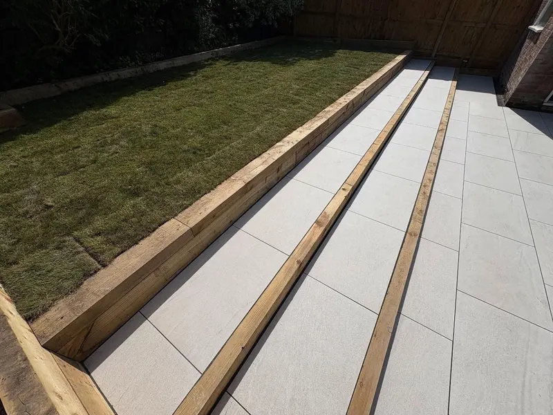 Garden tiers of light grey slabs divided by horizontal timber sleepers, with turf on the upper level and fencing around the plot.