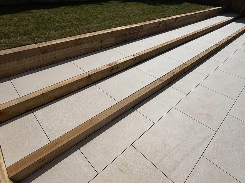 Angled detail of cream square paving on several levels, each step face formed by stacked timber sleepers below a green lawn.