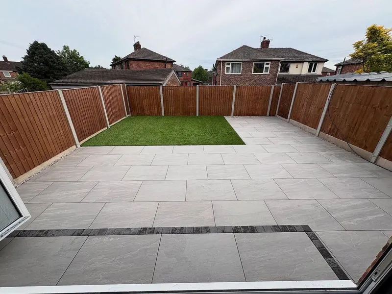 Fenced garden with staggered light grey paving, an artificial grass rectangle and a single row of dark bricks marking a layout change.
