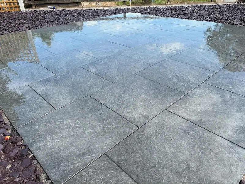 Wet light grey rectangular pavers in running bond beside a bed of dark purple decorative chippings, reflecting sky on the surface.