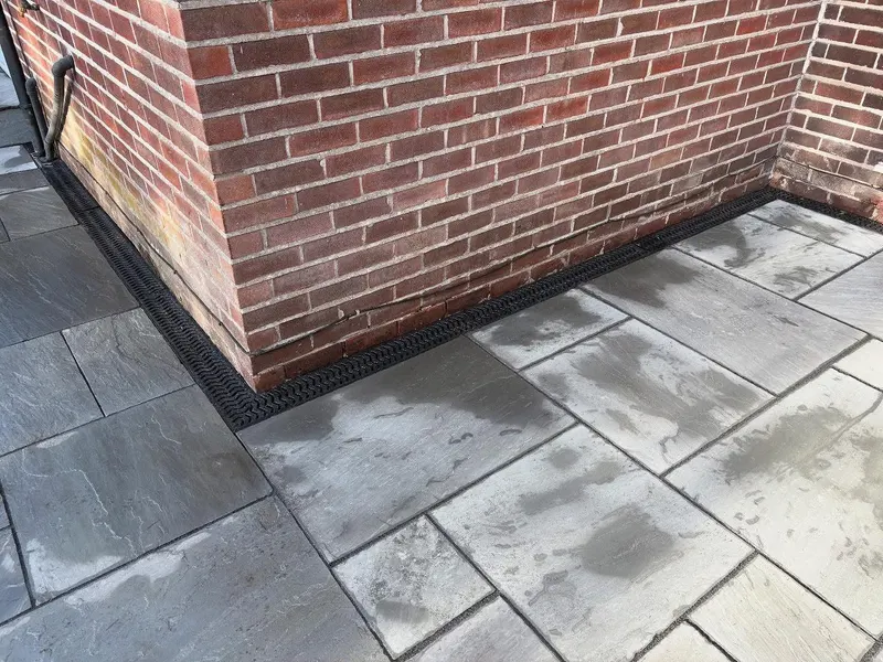 Mixed grey natural stone slabs along a brick wall corner, separated by a continuous black channel drain at the base of the masonry.