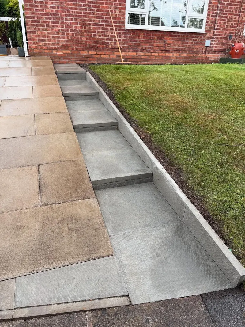 Flat buff-toned paving alongside six wide grey stone steps with a stone kerb, descending beside a sloped lawn to a brick wall.