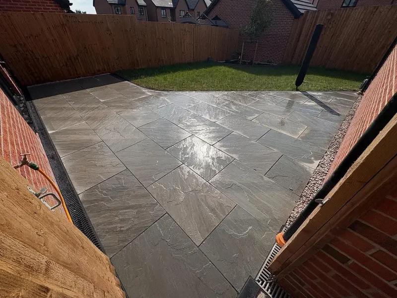 Riven stone slabs in grey and warm brown tones along a house wall, with a slot drain, gravel strip and green grass border.
