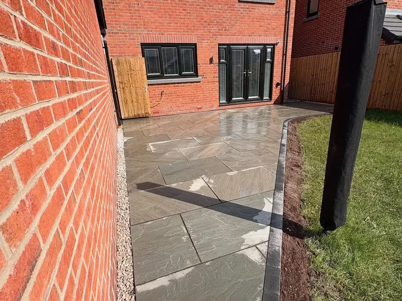 Grey natural stone flags sweeping around a lawn edge with a charcoal brick soldier course, gravel against the house wall and bright sun glare.