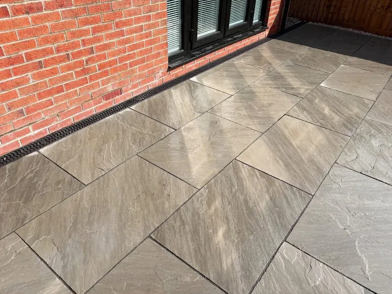 Rectangular riven slabs in grey and tan beside a red brick wall, with a black drainage channel and white gravel by a wooden fence.