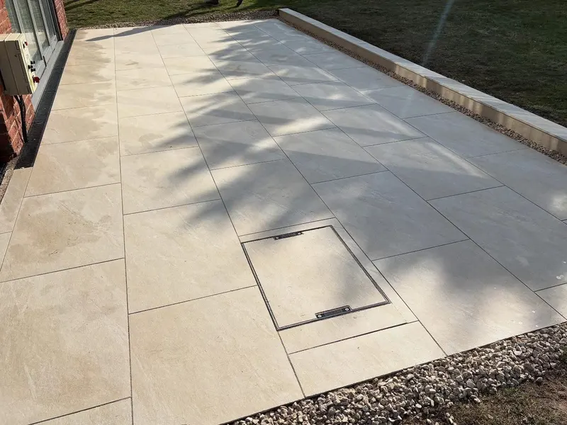 Smooth pale paving slabs with thin joints, a manhole cover set flush and infilled to blend, gravel edging and dappled tree shade.
