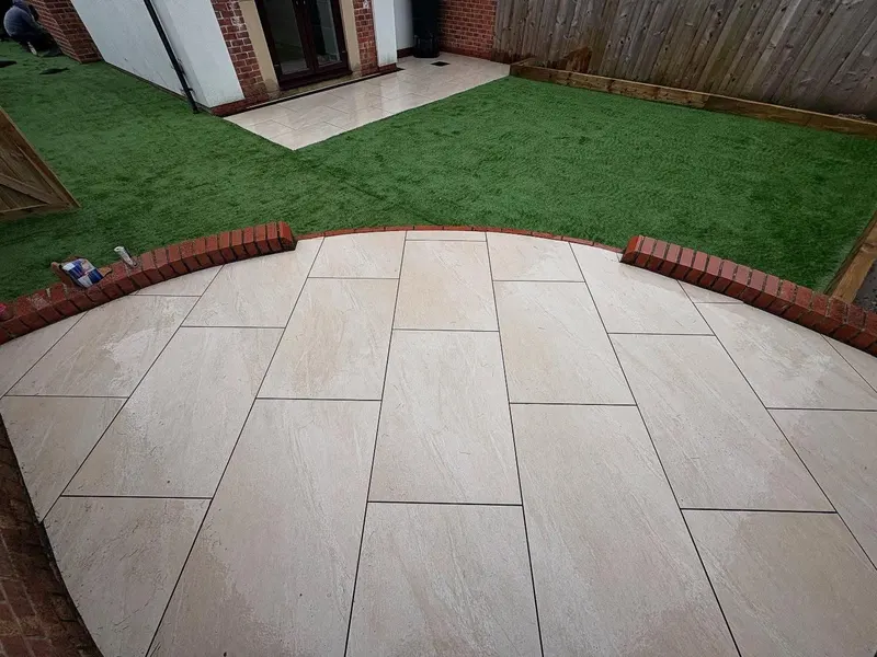 Curved section of cream rectangular paving bordered by red bricks on edge, opening onto artificial grass beside a brick house.