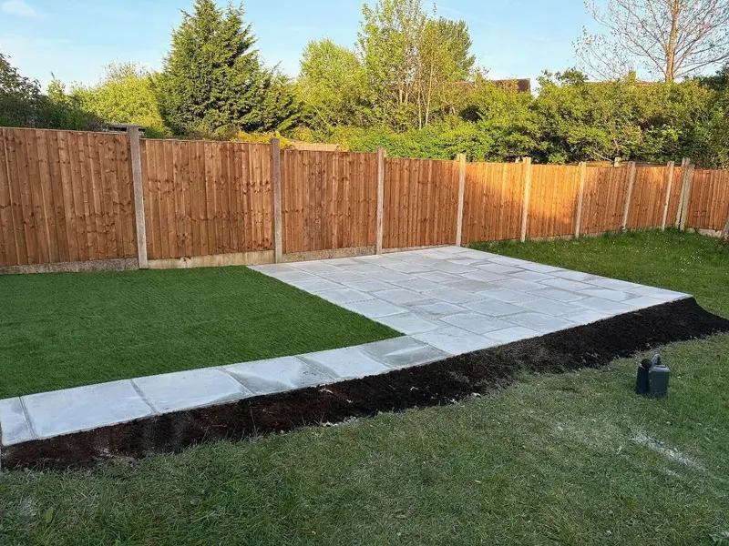 Light grey slab patio and a narrow paved strip running alongside a rectangle of artificial turf, with soil edging and a wooden fence.