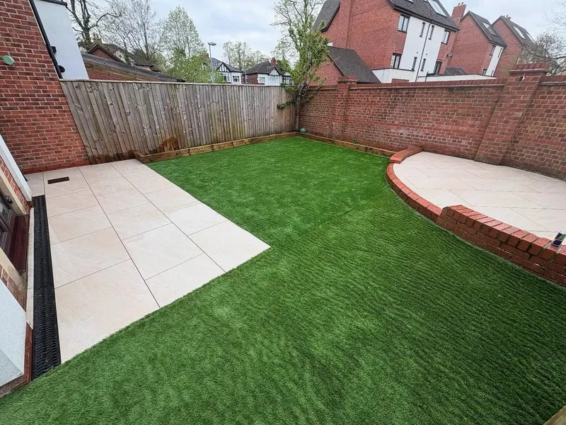 Back garden combining rectangular light paving by the house, a semicircular paved corner with low brick wall and artificial grass between.