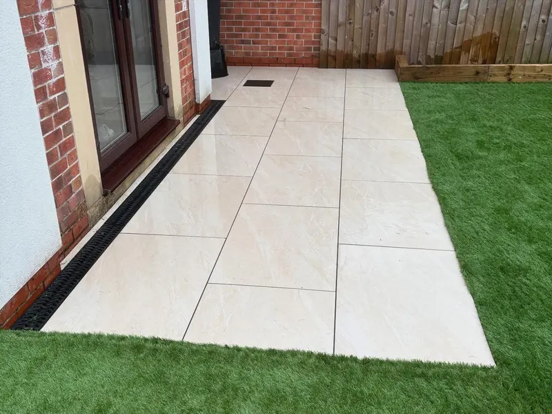 Large cream porcelain-style tiles in a grid beside neatly cut artificial grass, with channel drainage along the brick wall and timber bed beyond.