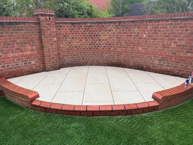 Fan-shaped light stone paving enclosed by a tall curved red brick garden wall and a low brick kerb facing a green lawn.