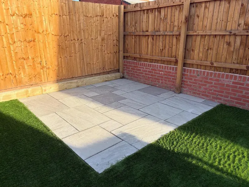 Light mixed-size stone flags filling a fenced corner next to artificial grass, with timber and brick boundary walls casting sharp shadows.