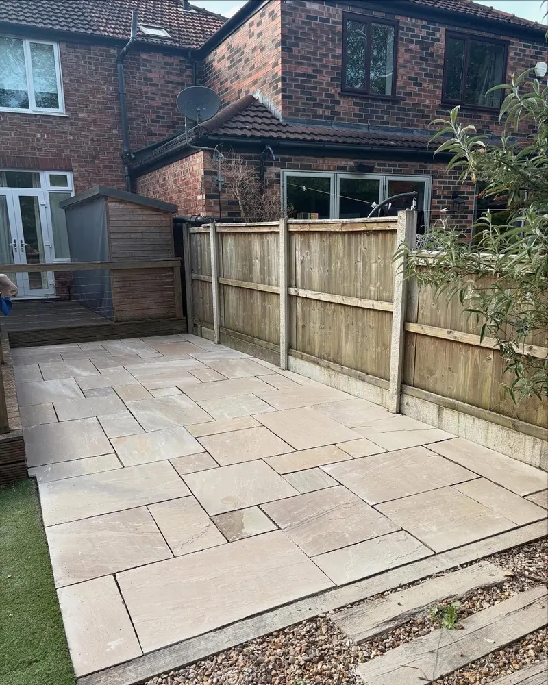 Random-pattern buff and cream stone paving stretching past a wooden fence toward a red brick house, with gravel and sleeper edging in front.
